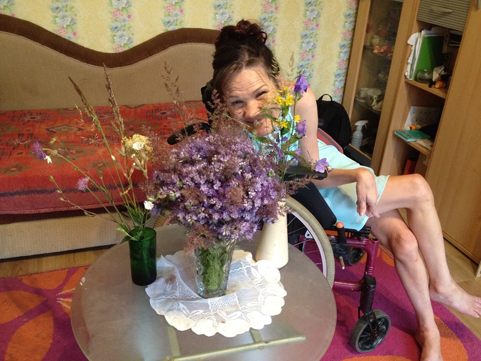 Naeratav ja ratastoolis istuv tumepruunide juustega naine lillakate lillede taamal enda kodus. 